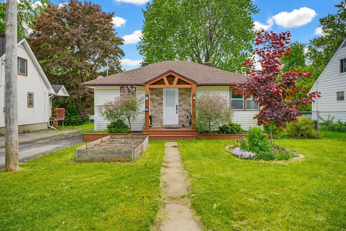 Single-story white house with stone entryway, wooden porch, front garden, and a vibrant red maple tree on a sunny day.