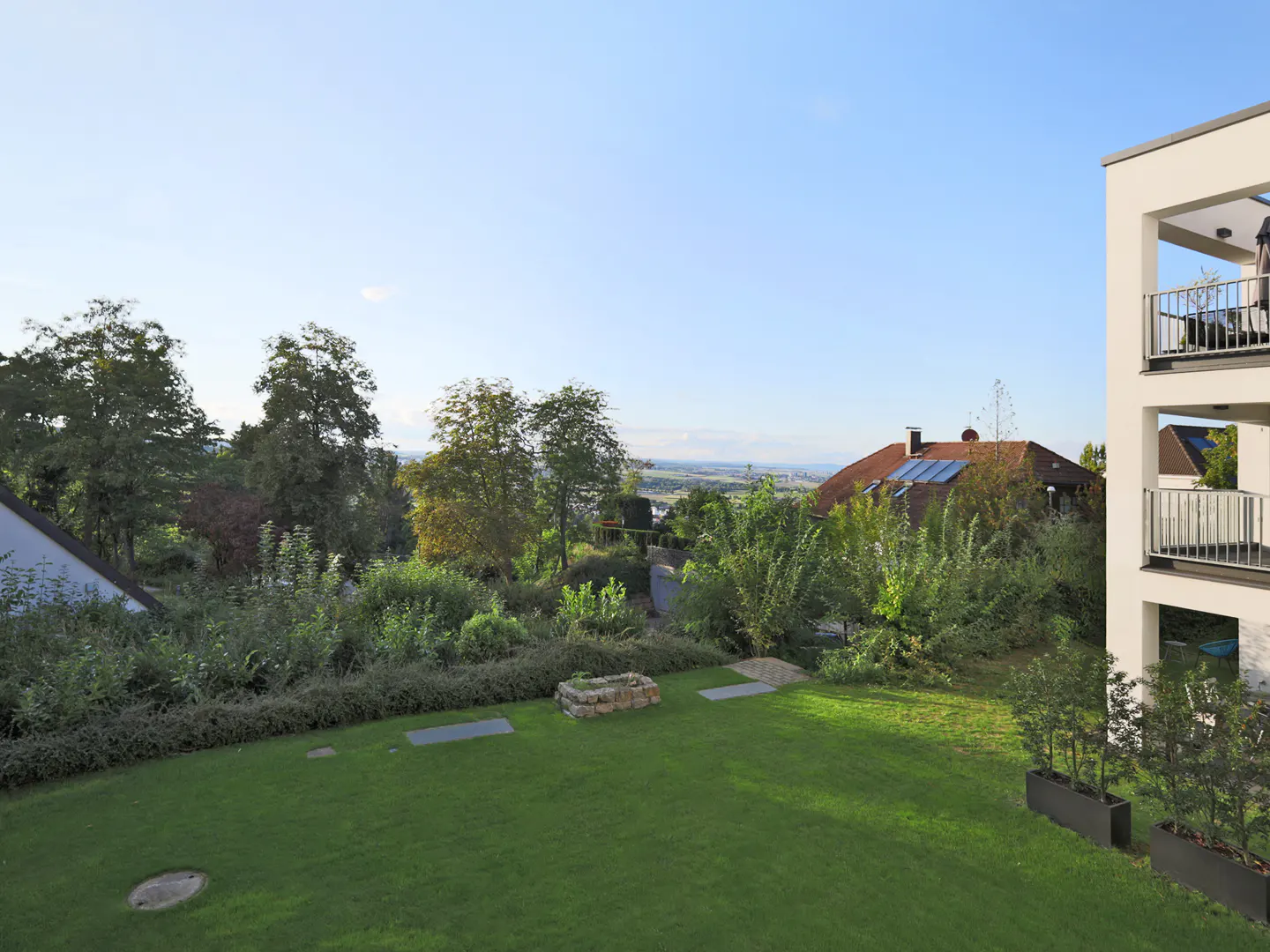 View of a green lawn with trees, a house with a red roof, and a modern white building with balconies under a blue sky.