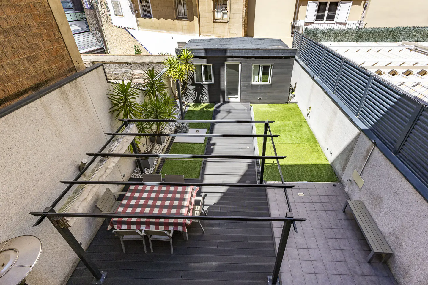 Overhead shot of a modern backyard with a dining table under a pergola, a small gray house, and artificial grass.
