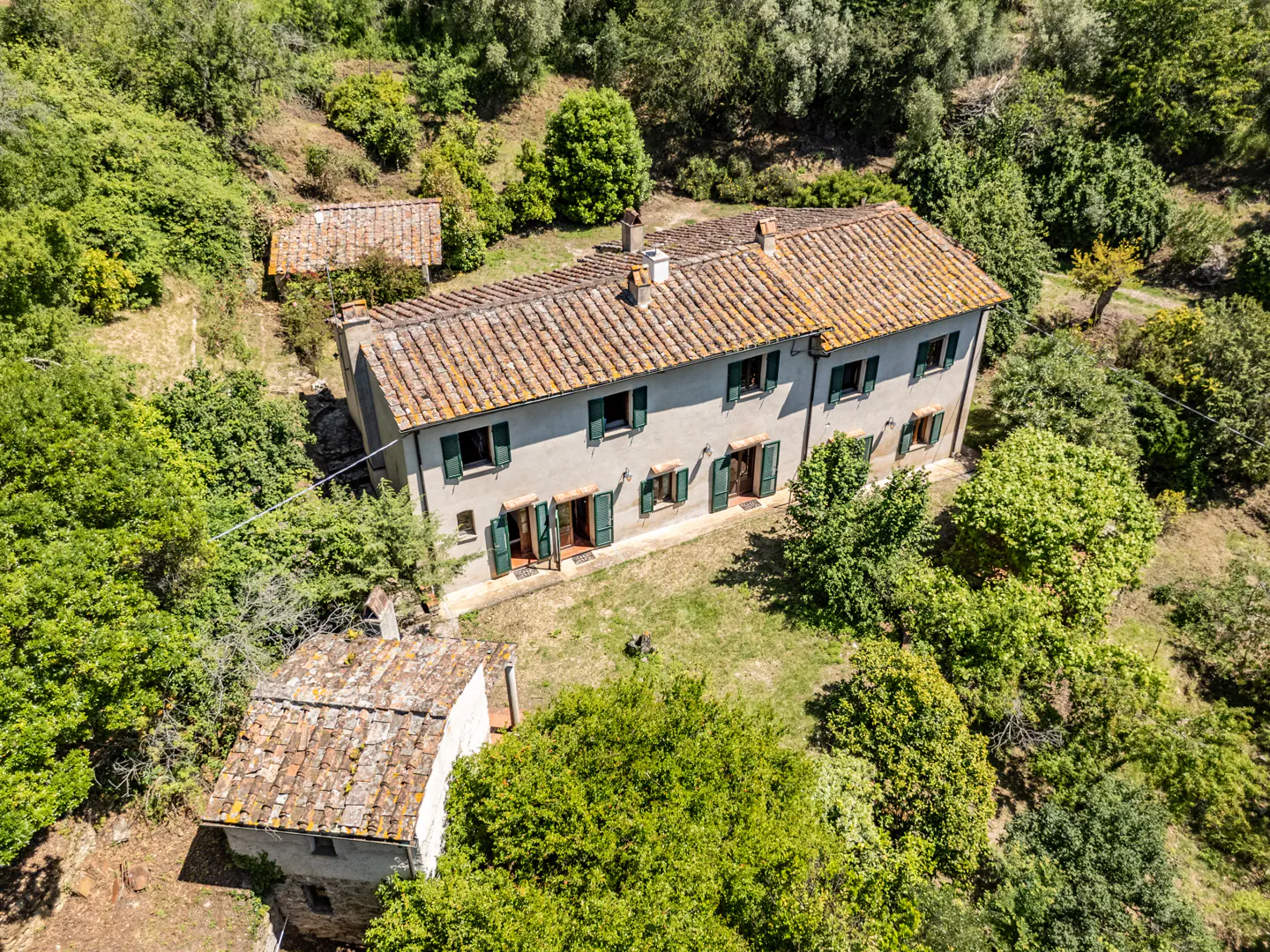 Aerial view of a two-story house with a terracotta tile roof and green shutters, surrounded by lush green trees and a smaller outbuilding.