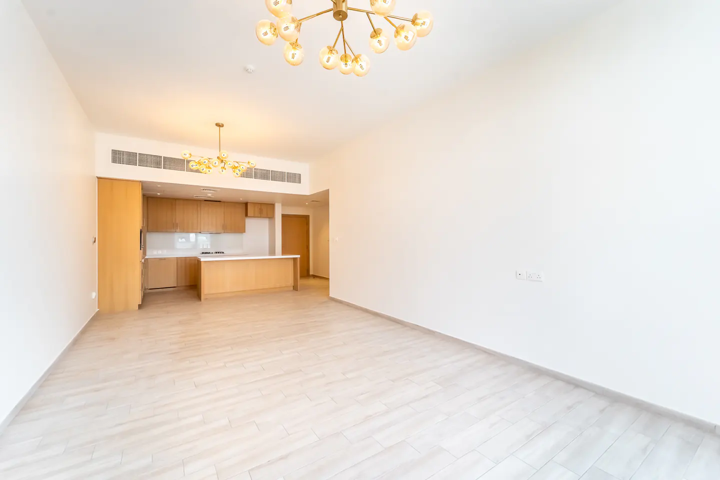 Bright, empty apartment with light wood floors, white walls, and a modern chandelier. The kitchen has light wood cabinets and a white countertop.