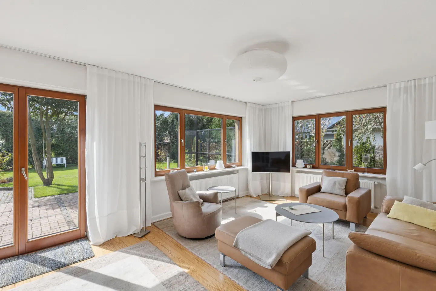 Bright living room with brown leather furniture, wood floors, and large windows overlooking a green yard. White curtains frame the windows.