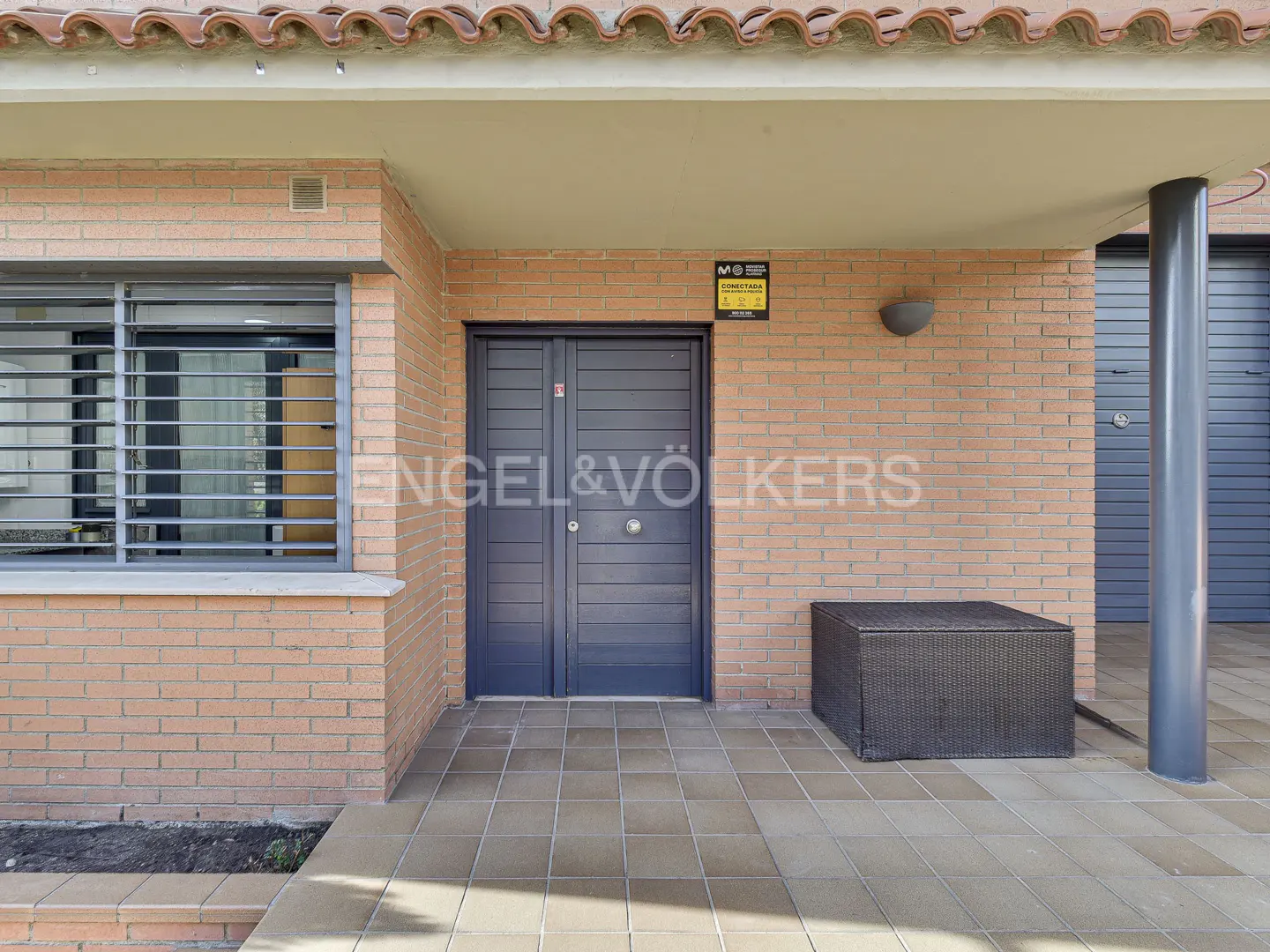 Exterior view of a brick building with a gray double door, window with bars, and a dark wicker bench. The Engel & Völkers logo is visible.