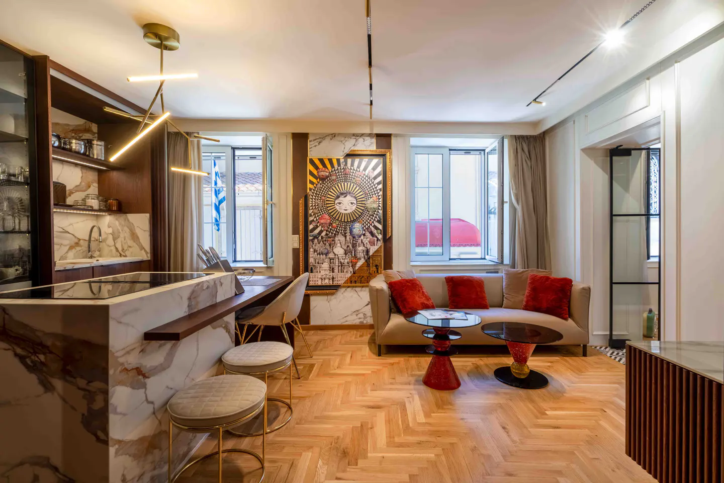 Modern open-plan living area with marble kitchen island, bar stools, beige sofa with red pillows, and unique wall art.