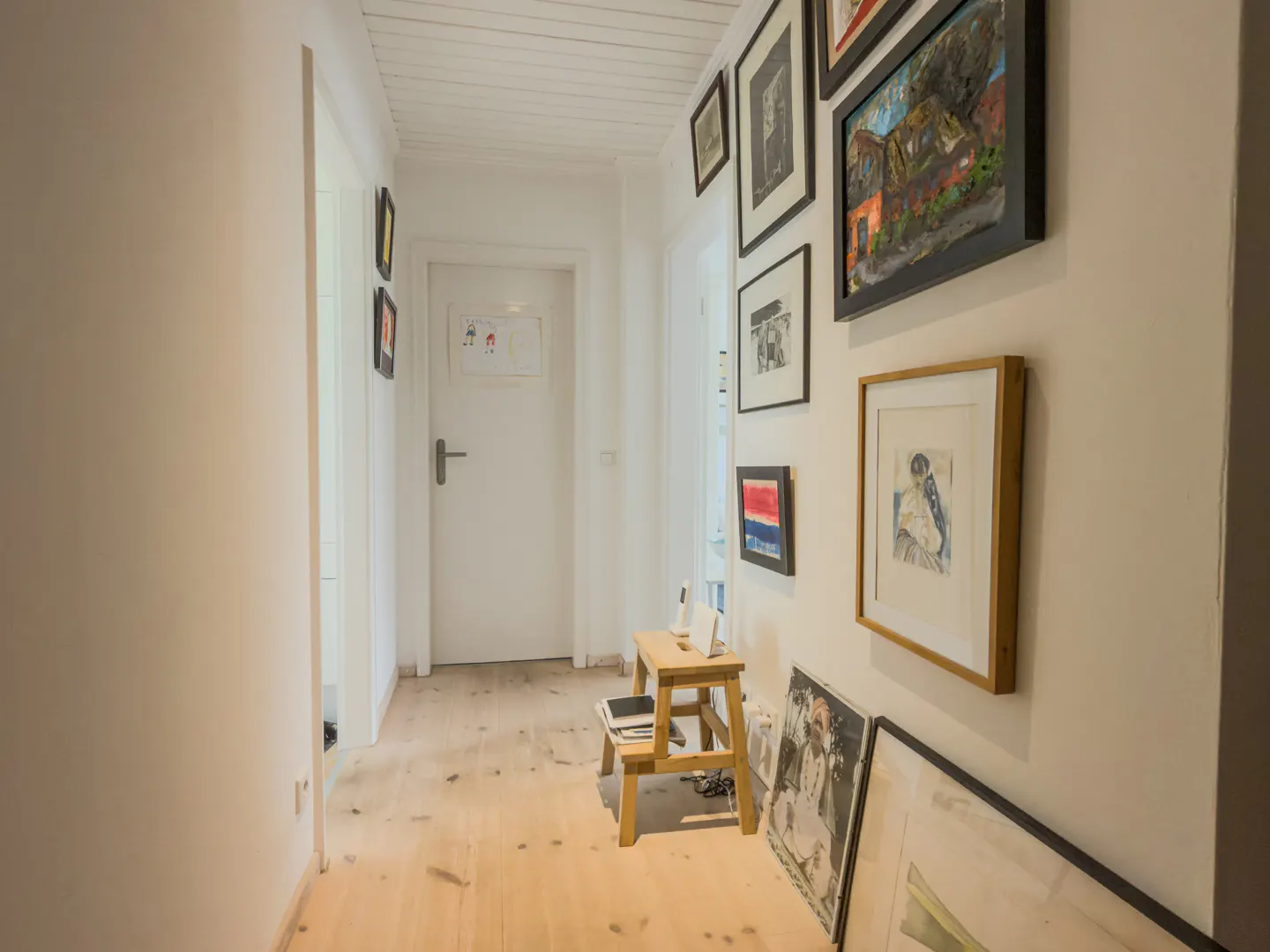 Hallway with white walls, light wood floors, and a gallery wall of framed art. A small wooden step stool sits near the wall.