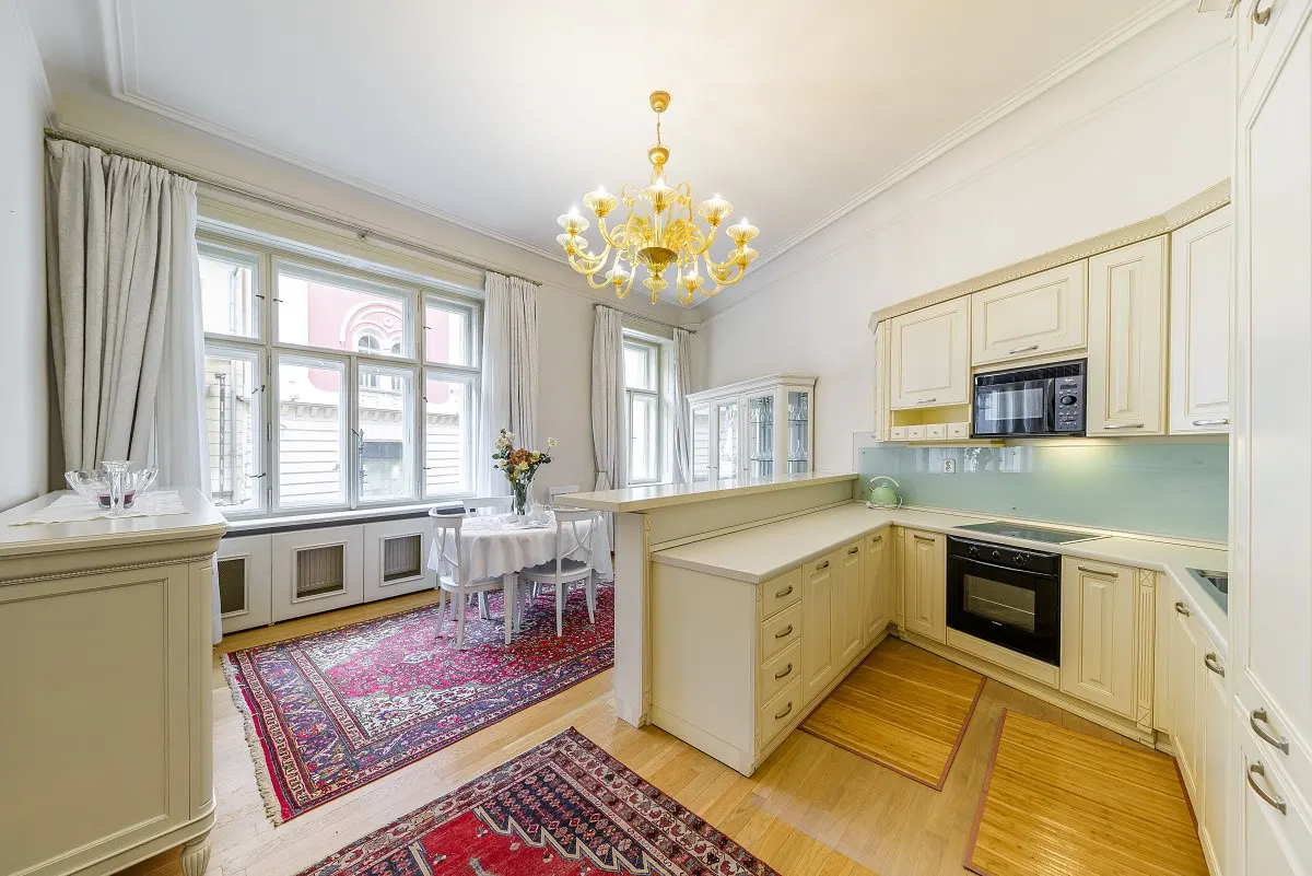 Bright kitchen with white cabinets, a gold chandelier, and a dining area with a red rug. Large windows provide natural light.