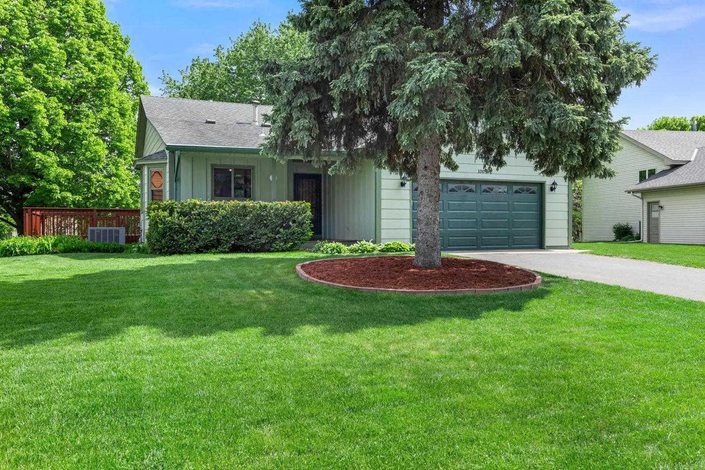 A light green house with a dark green garage door and a large tree in the front yard.