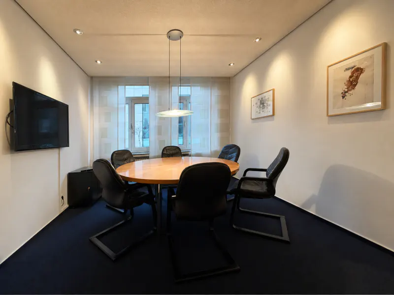 Conference room with a round wood table, black chairs, and a flat-screen TV on the wall. Art hangs on the walls, and natural light filters through sheer curtains.