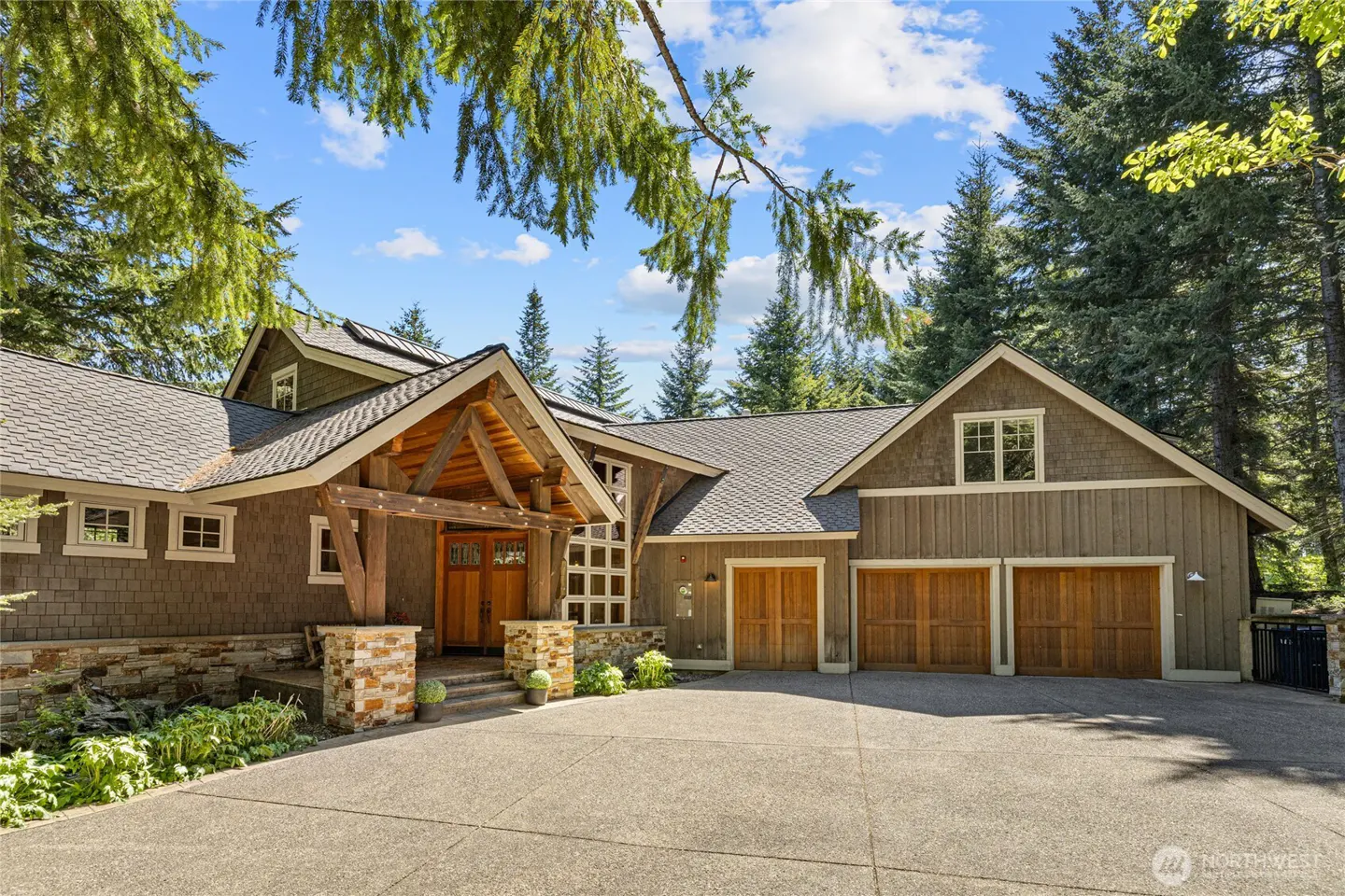 A rustic brown home with a stone-accented entryway, wooden garage doors, and a concrete driveway surrounded by tall trees.