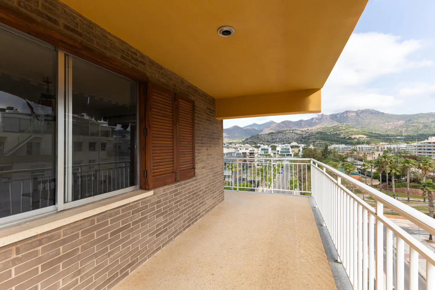 Balcony view with brick wall, brown shutters, and white railing overlooking a city with mountains in the background.