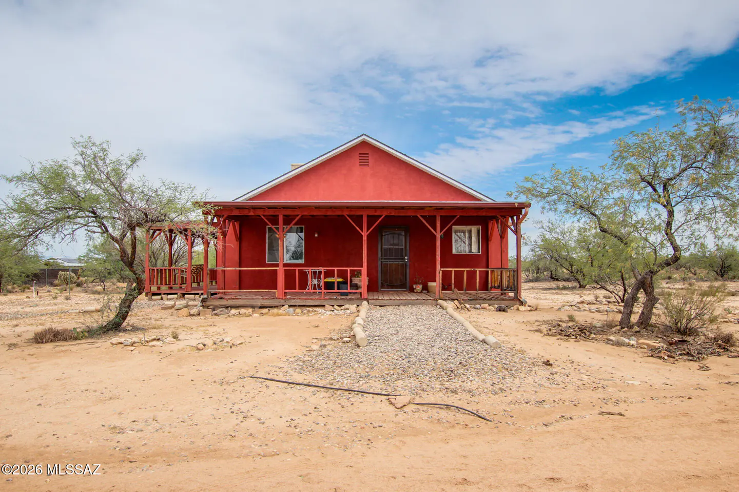 Red, single-story house with a covered porch and a gravel path in a desert landscape under a blue sky.