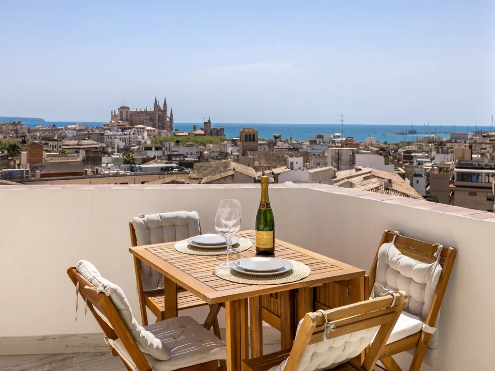 Rooftop dining with city view. Table set with champagne, plates, and glasses. Cathedral and ocean in background.