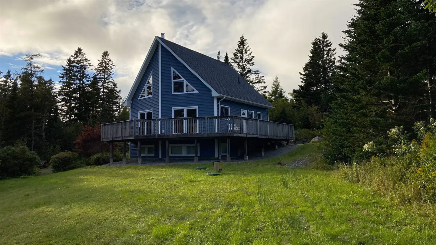 Blue A-frame house with a deck, surrounded by trees and a grassy lawn.