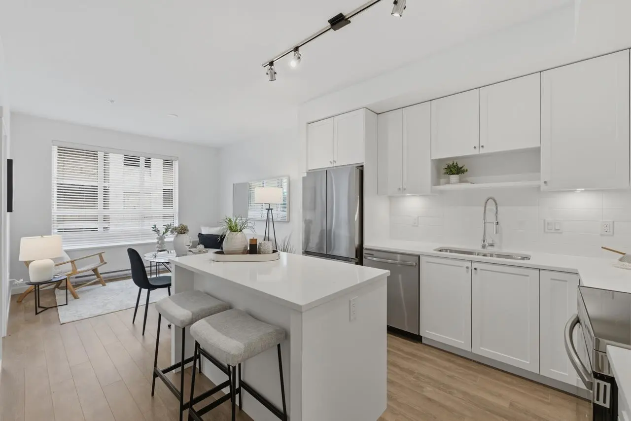 Bright, modern condo with white kitchen cabinets, stainless steel appliances, and a quartz island with two stools. Living area with a chair and table.