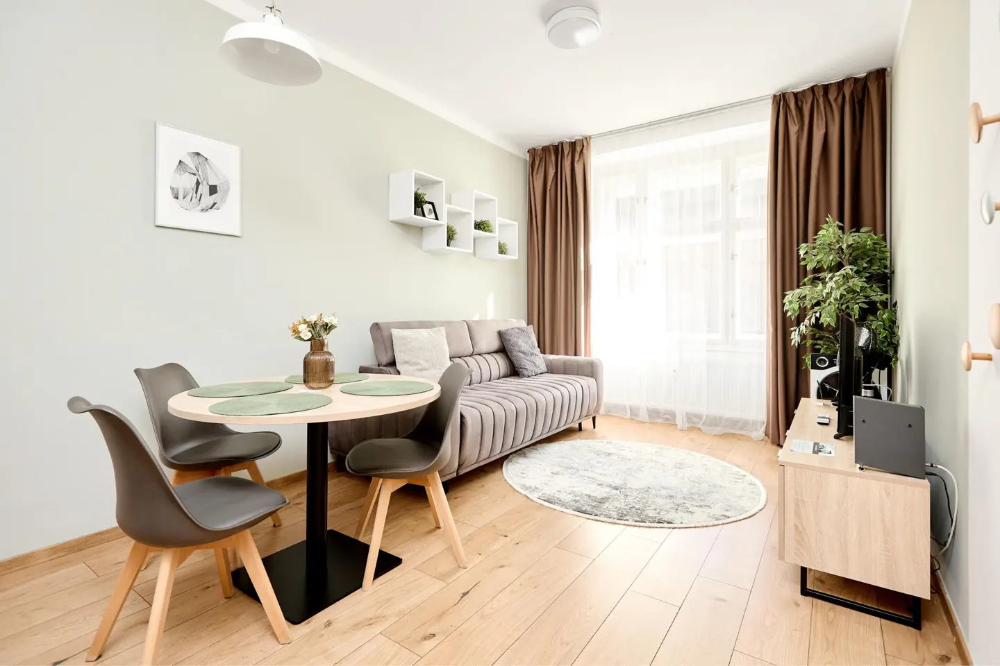 A bright living room with a round table, four chairs, a sofa, and a TV stand. Brown curtains frame a large window.
