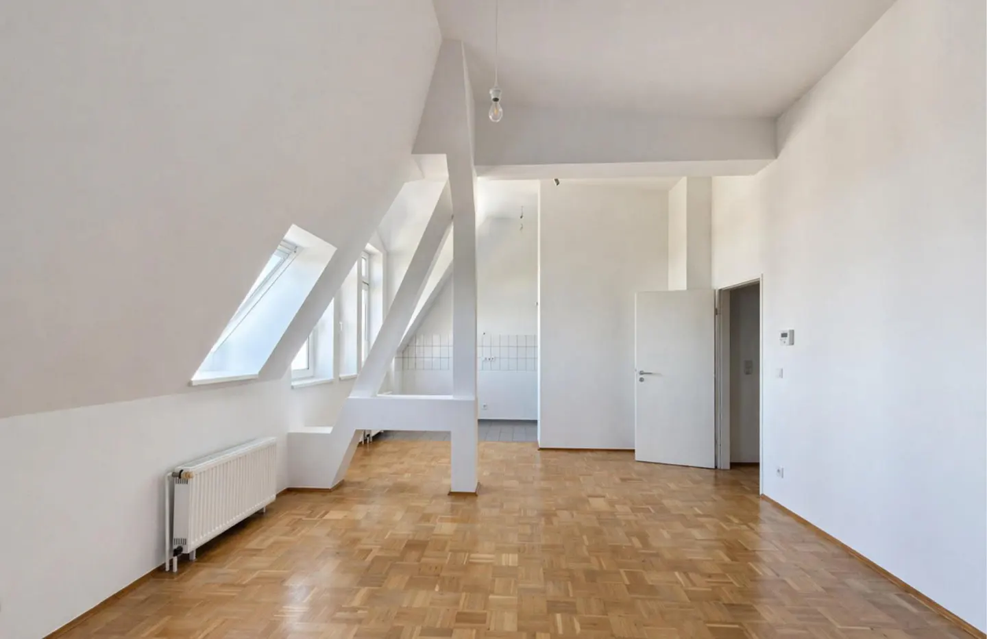 Bright, empty attic apartment with white walls, parquet floor, skylights, and a view into the kitchen.