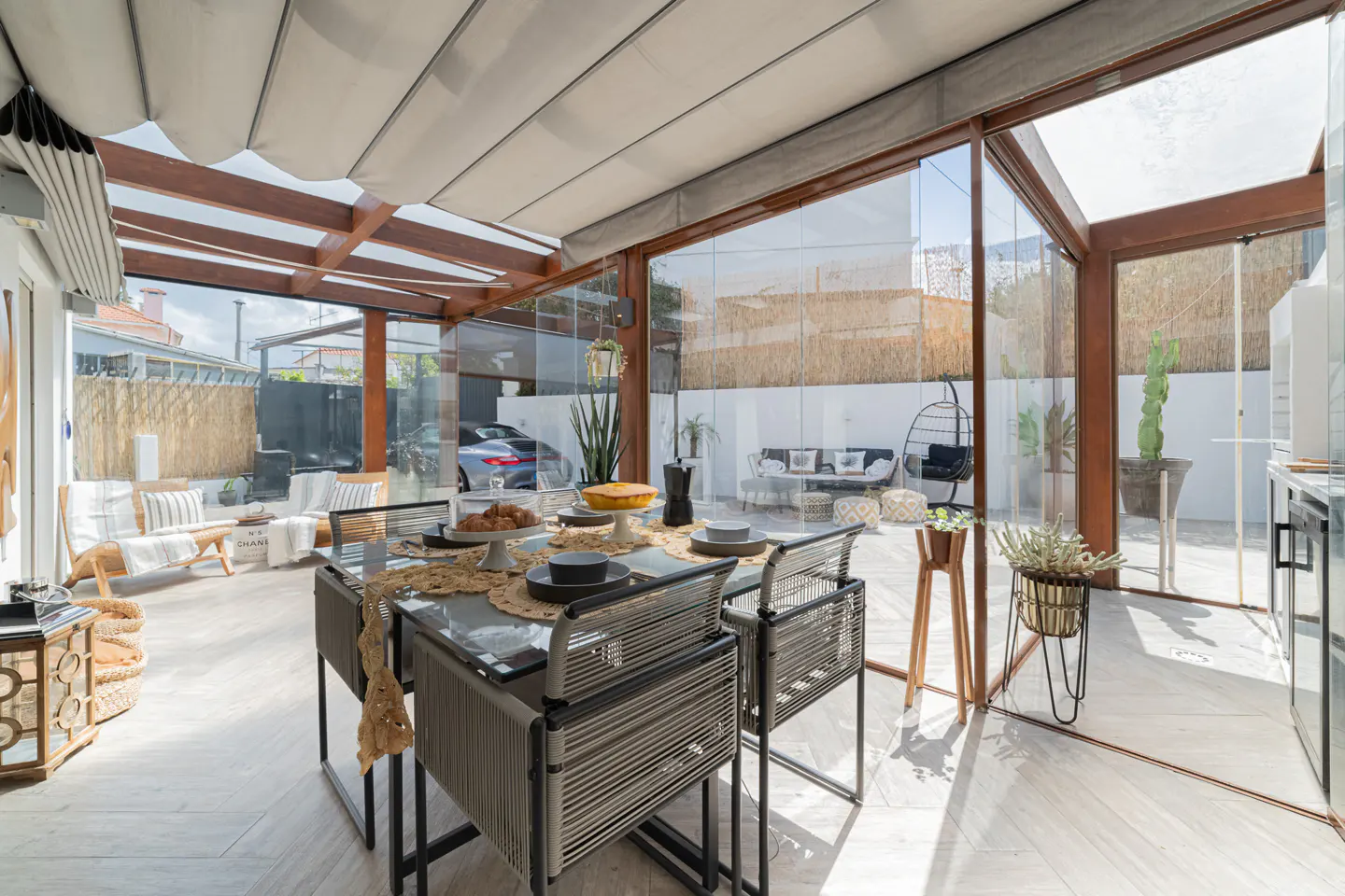 Bright sunroom with a glass-enclosed dining area, a table set with food, and a lounge area visible through the glass.