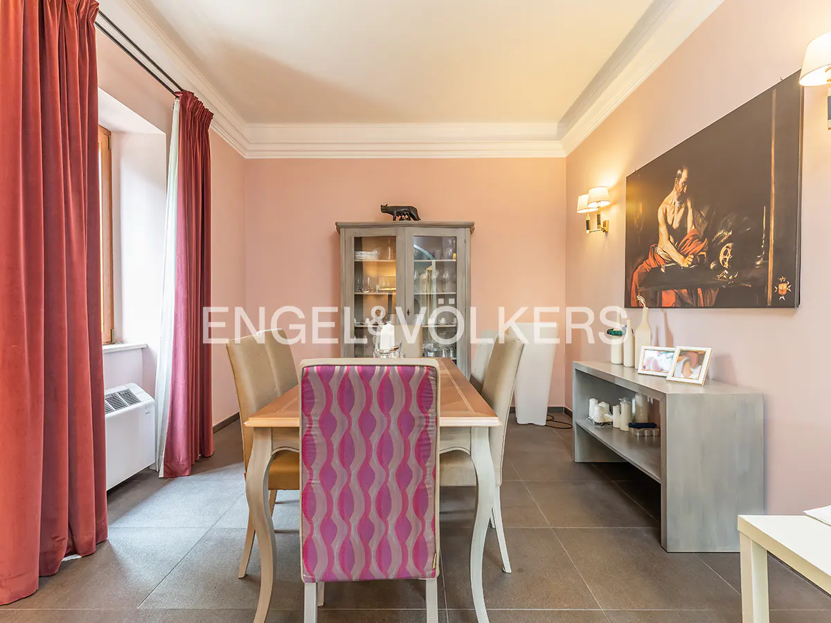 A dining room with pink walls, a wooden table with four chairs, and a gray cabinet with glass doors. A large painting hangs on the wall.