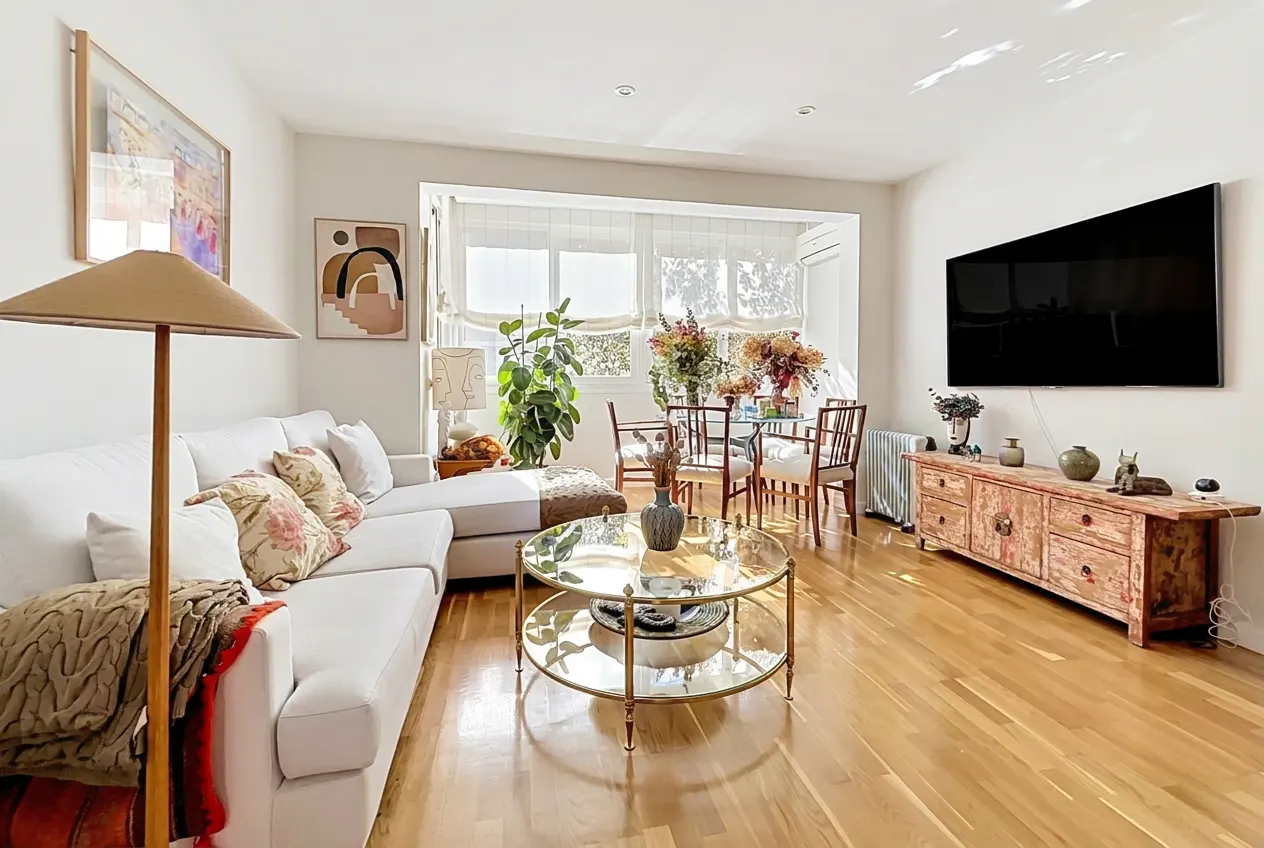 Bright living room with a white sofa, round glass table, and a dining area by the window. A large TV hangs above a distressed wooden cabinet.