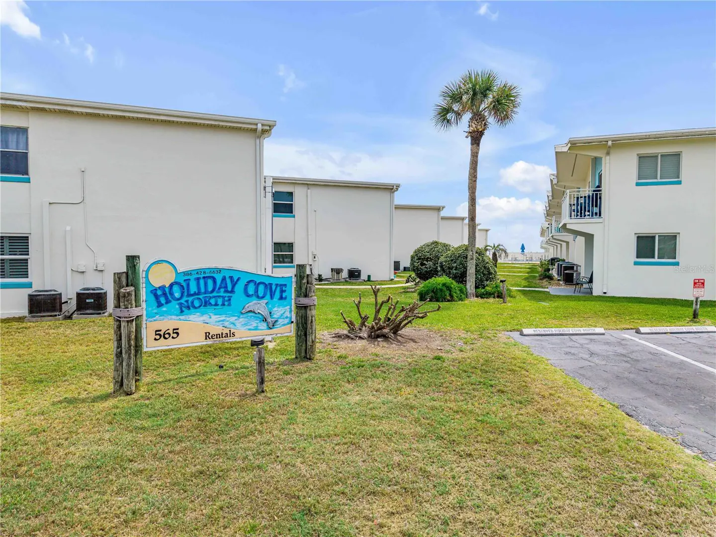 Holiday Cove North rental condos with a blue sign, palm tree, and green lawn under a partly cloudy sky.