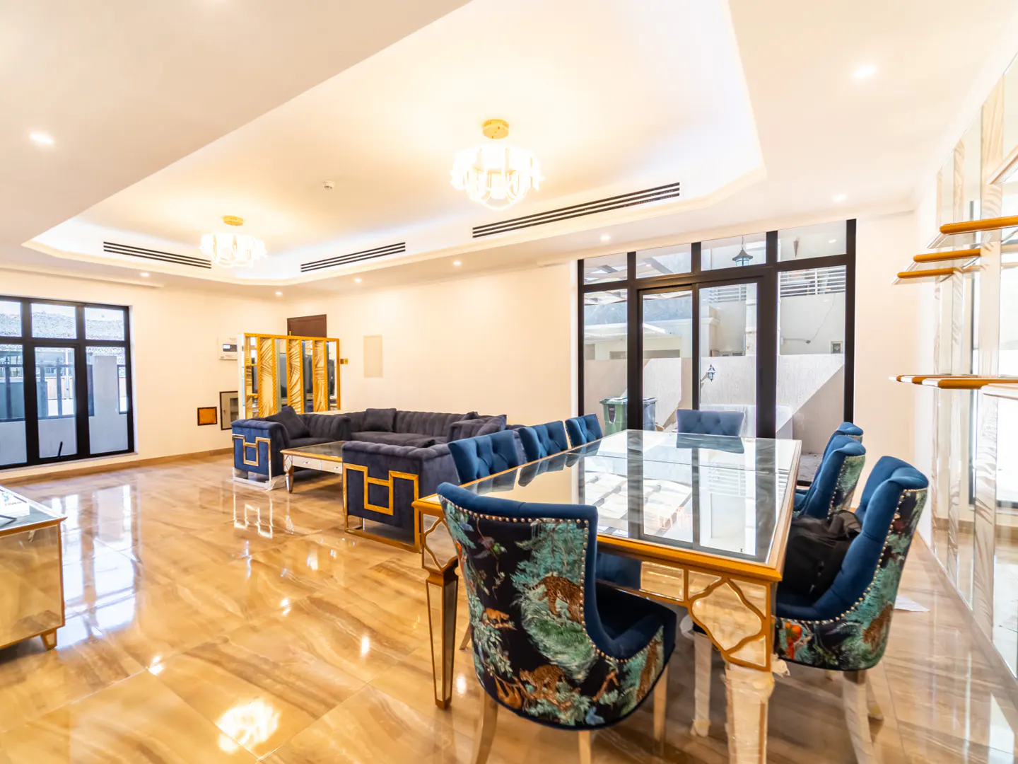 Modern living room with a glass dining table, blue patterned chairs, and a matching blue sectional sofa on marble floors.