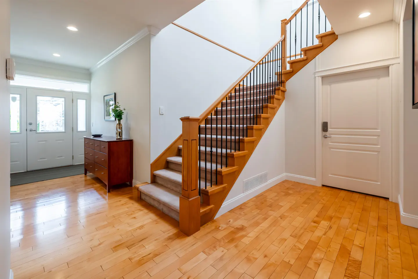 Bright foyer with hardwood floors, a staircase with black railings, and a white front door with glass panels. A brown dresser sits near the entrance.