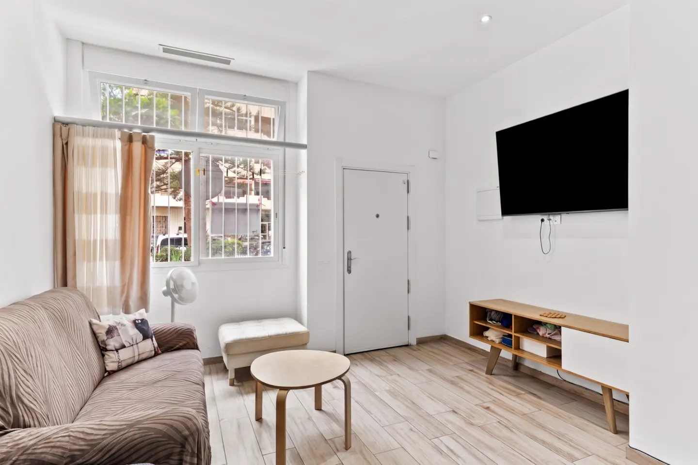 Living room with a brown sofa, round table, TV, and white walls. A window with curtains lets in natural light.