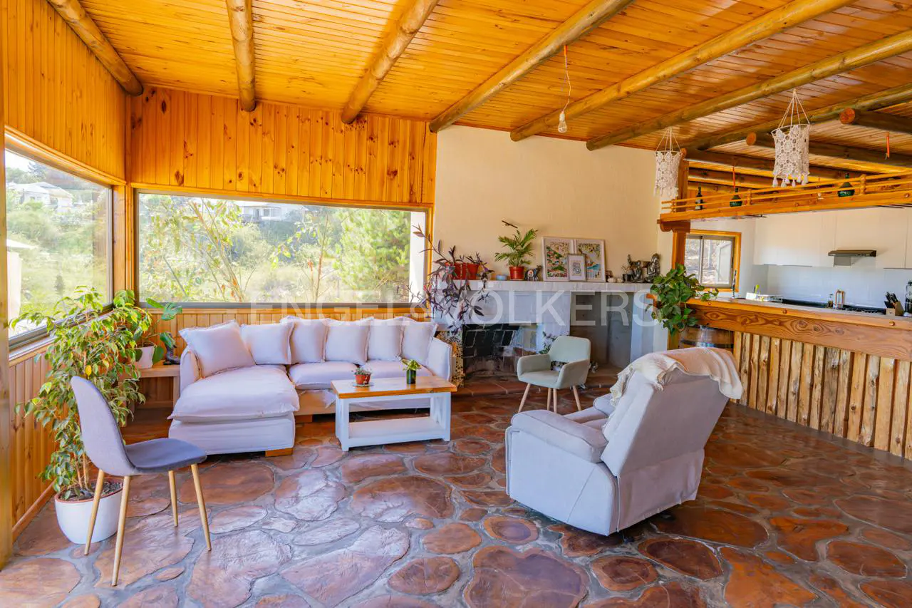 A cozy living room with wood-paneled walls, a stone floor, and a white sectional sofa. A large window offers a view of greenery.