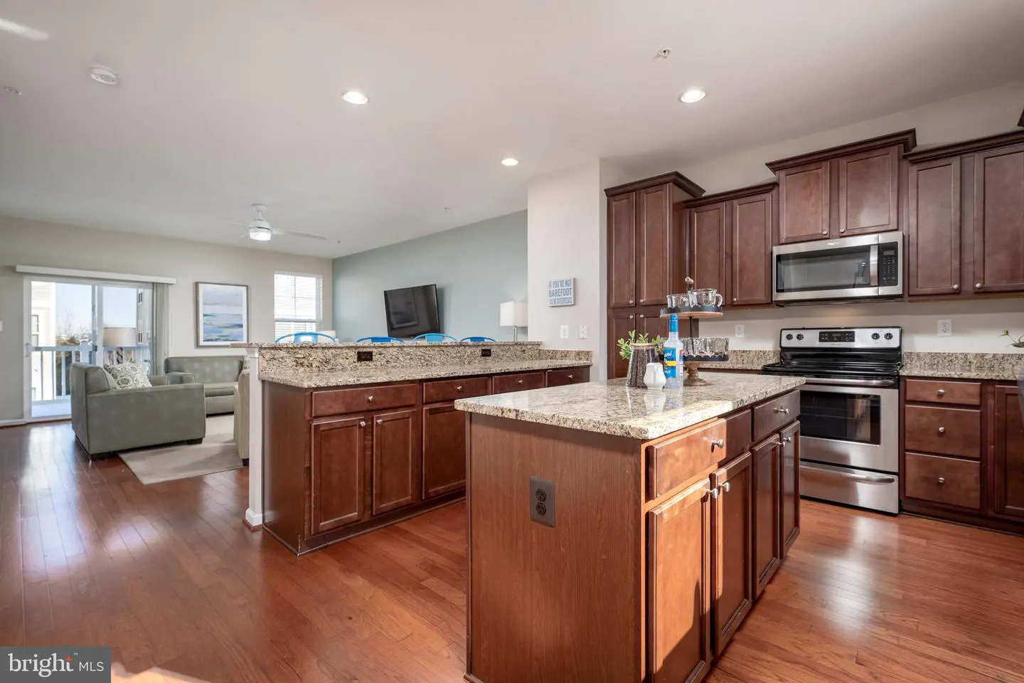 Open concept kitchen with dark wood cabinets, granite countertops, stainless steel appliances, and hardwood floors. Living room in background.
