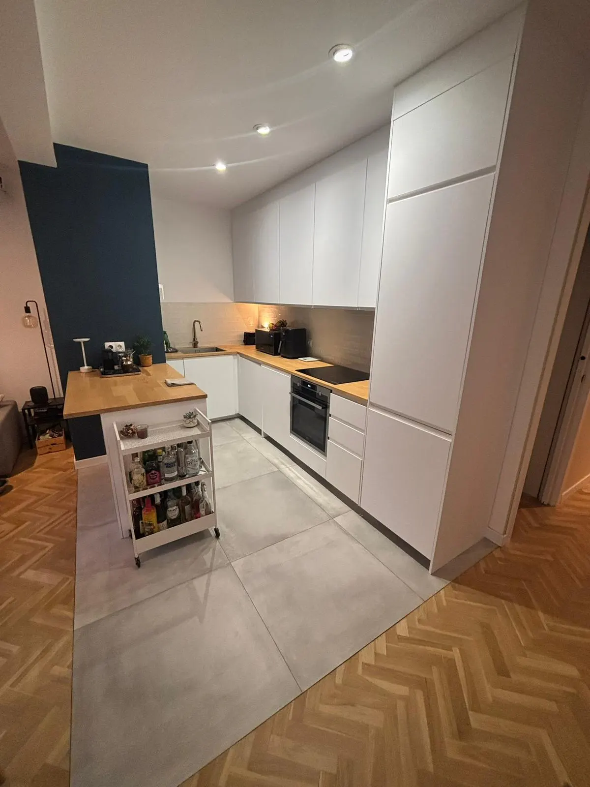 Bright kitchen with white cabinets, wood countertops, and stainless steel appliances. A rolling cart holds bottles. Herringbone wood floor.