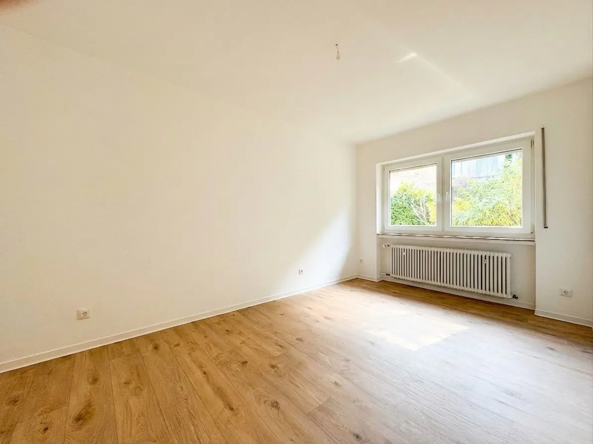 Bright, empty room with wood floors, white walls, and a window showing green foliage. A white radiator sits beneath the window.