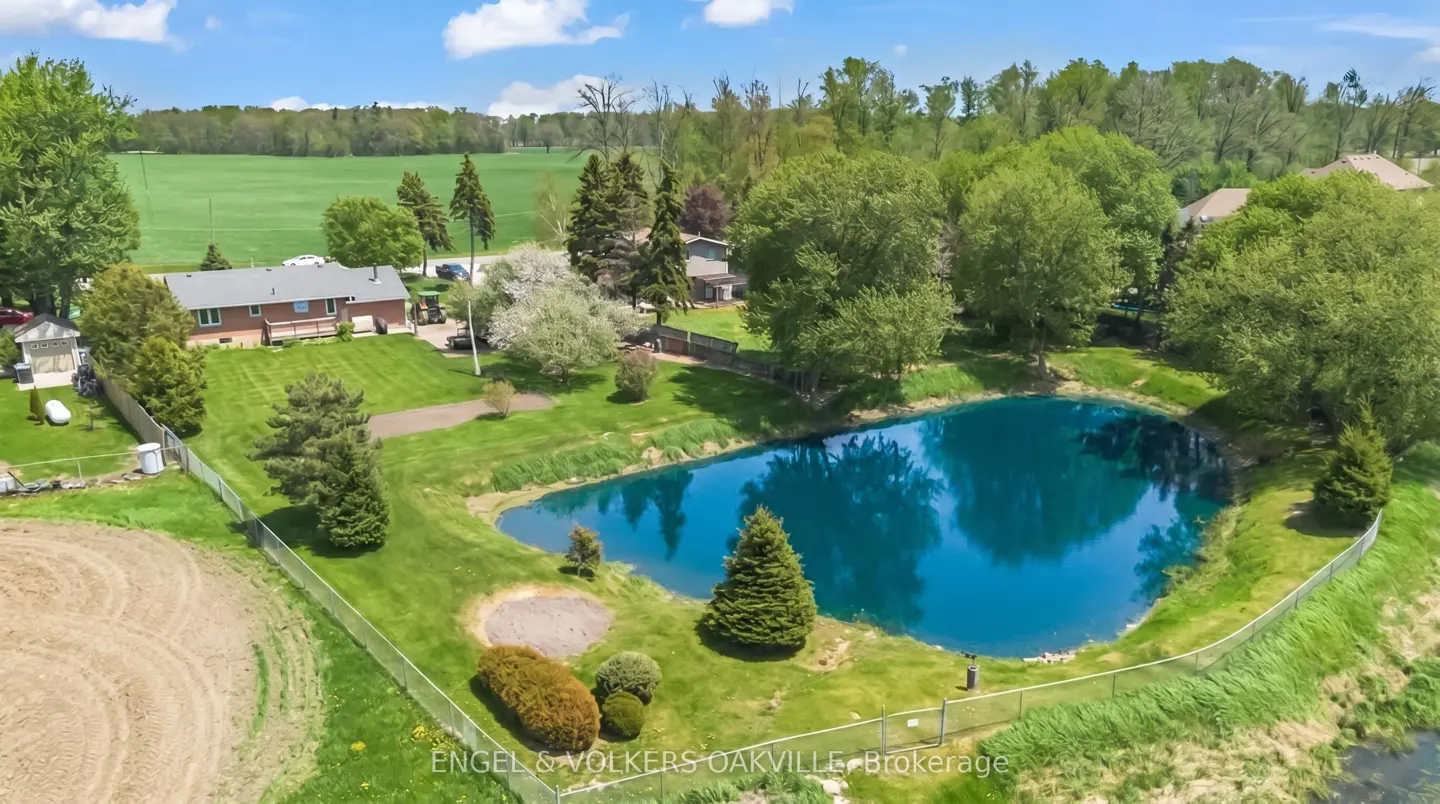 Aerial view of a red brick house with a blue pond, green grass, and trees under a blue sky.