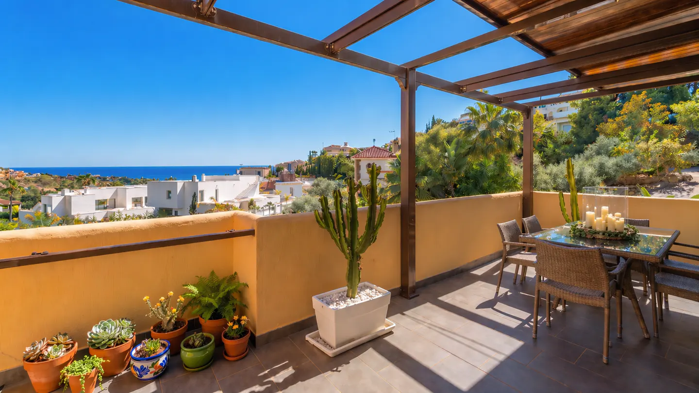 A sunny patio with a table, chairs, and potted plants overlooks white buildings and the ocean under a blue sky.