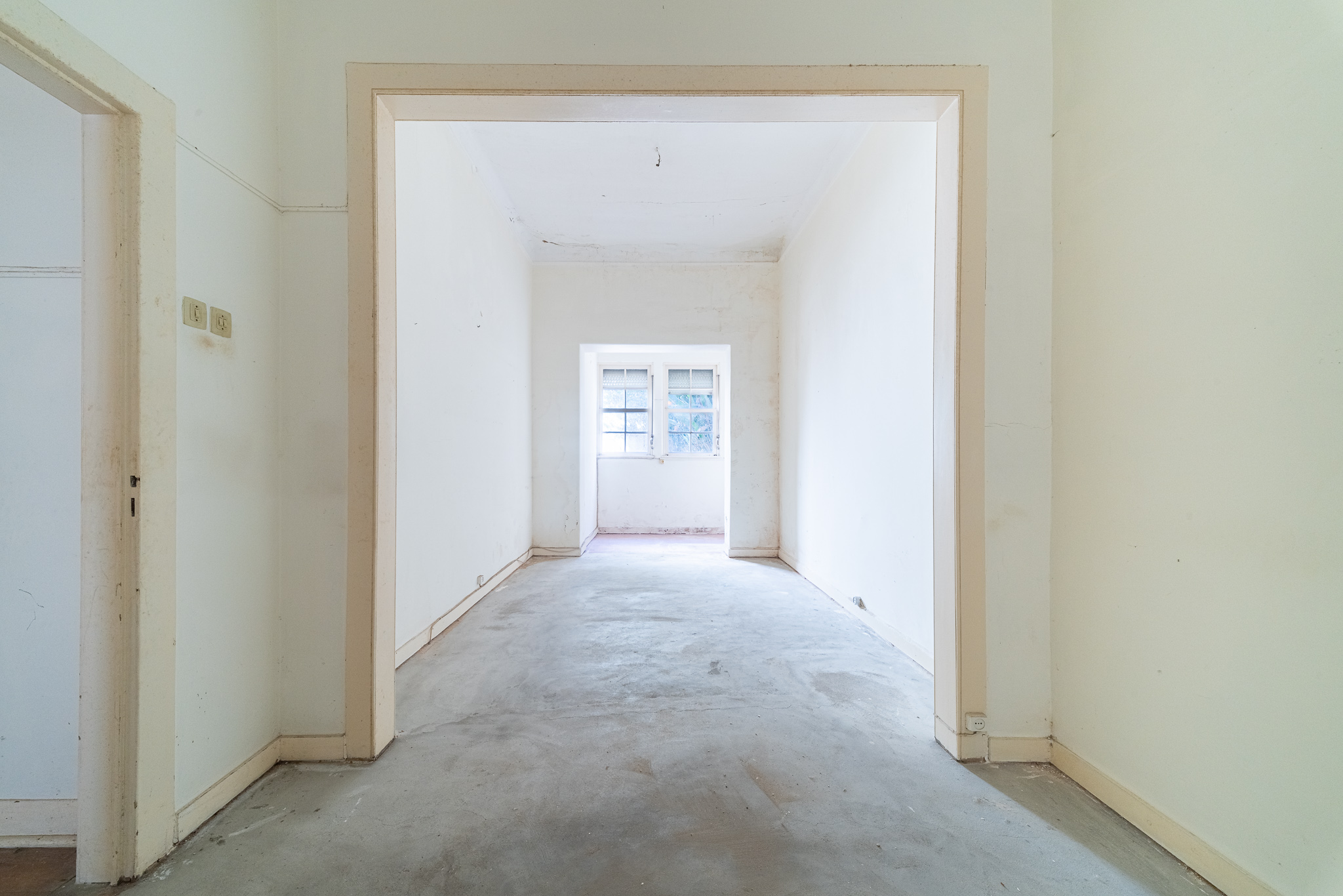 Empty, unfurnished room with white walls, concrete floors, and a doorway leading to a bright window at the far end. [AI-generated text]