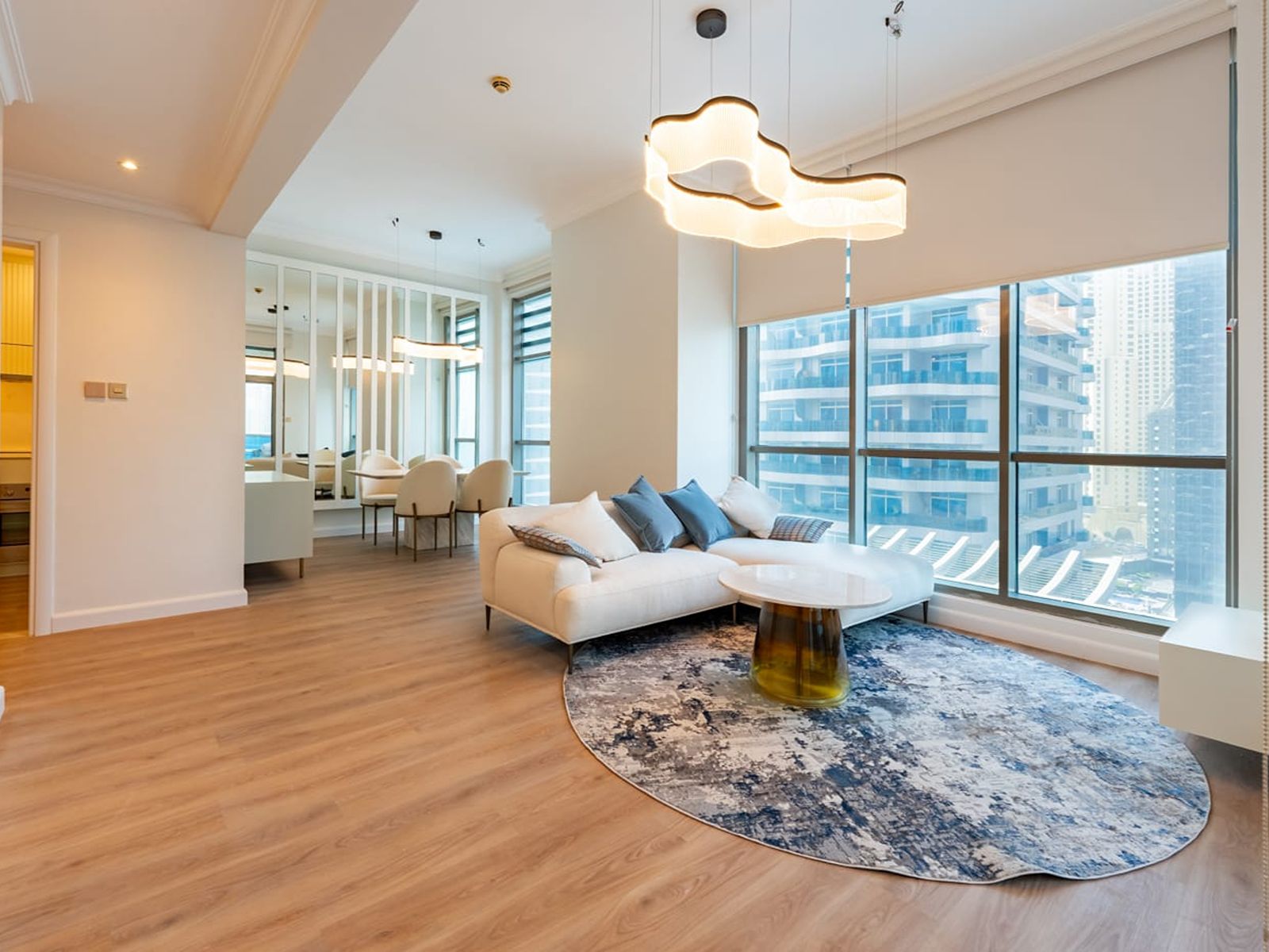 Modern living room with white sofa, blue pillows, round patterned rug, and large windows overlooking city buildings. [AI-generated text]