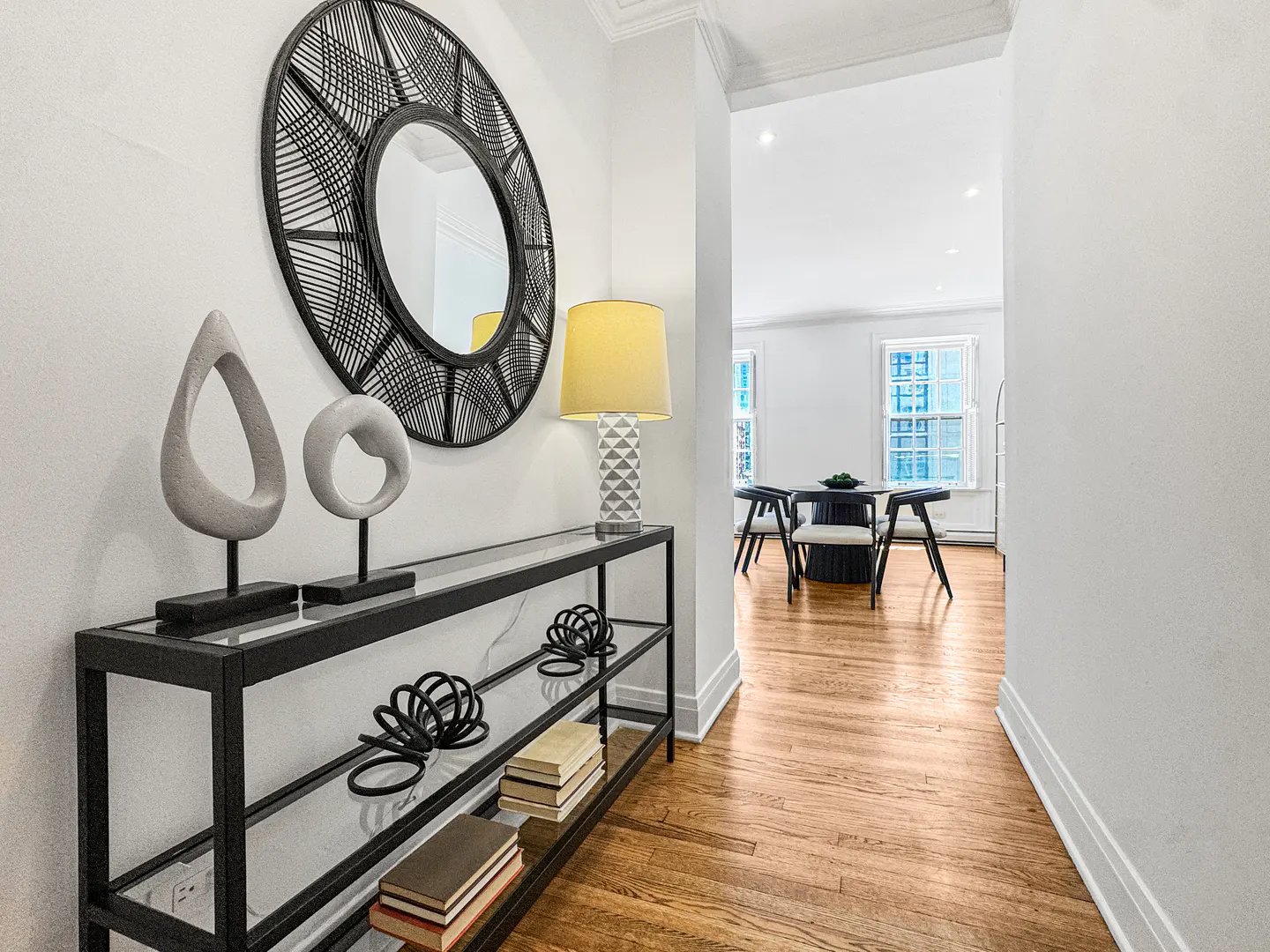 Entryway with a black console table, round mirror, lamp, and sculptures against a white wall. A dining area is visible in the background.