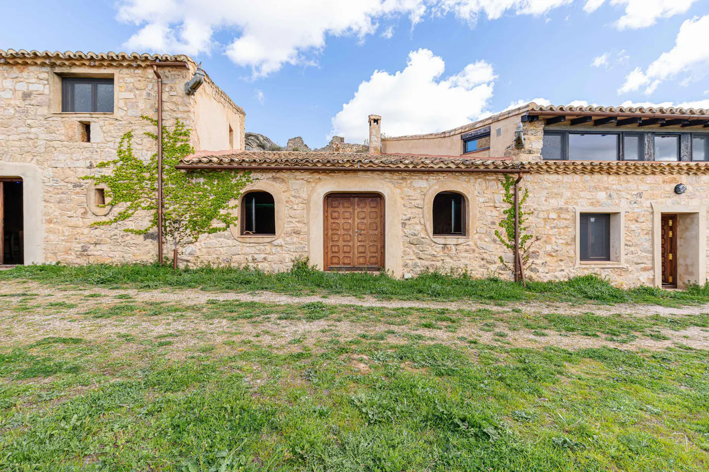 Exterior view of a stone house with a brown wooden door, arched windows, and green vines against a blue sky.