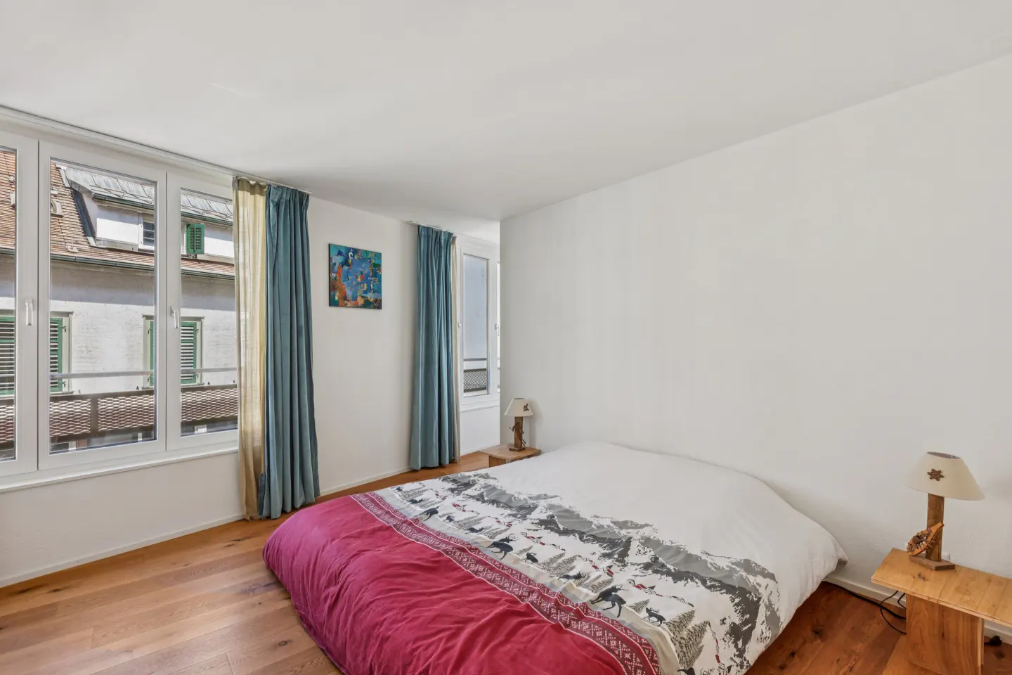 Bedroom with wood floors, white walls, and a bed with a red and white patterned duvet. Blue curtains frame a window with a view of buildings.