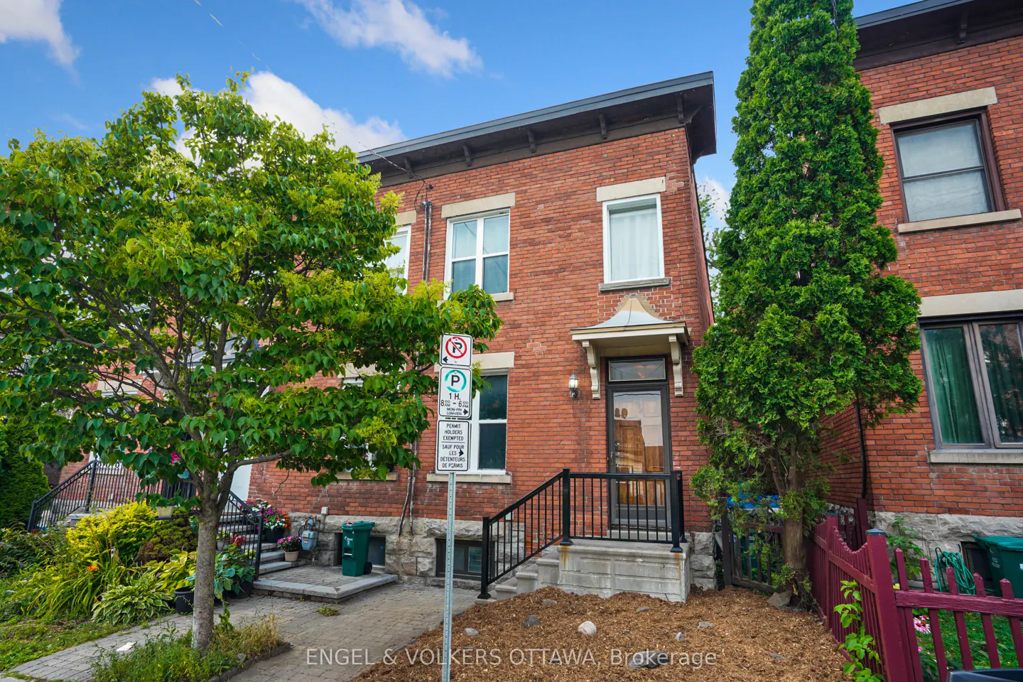 Exterior view of a two-story red brick house with black railings, a green tree, and a blue sky.