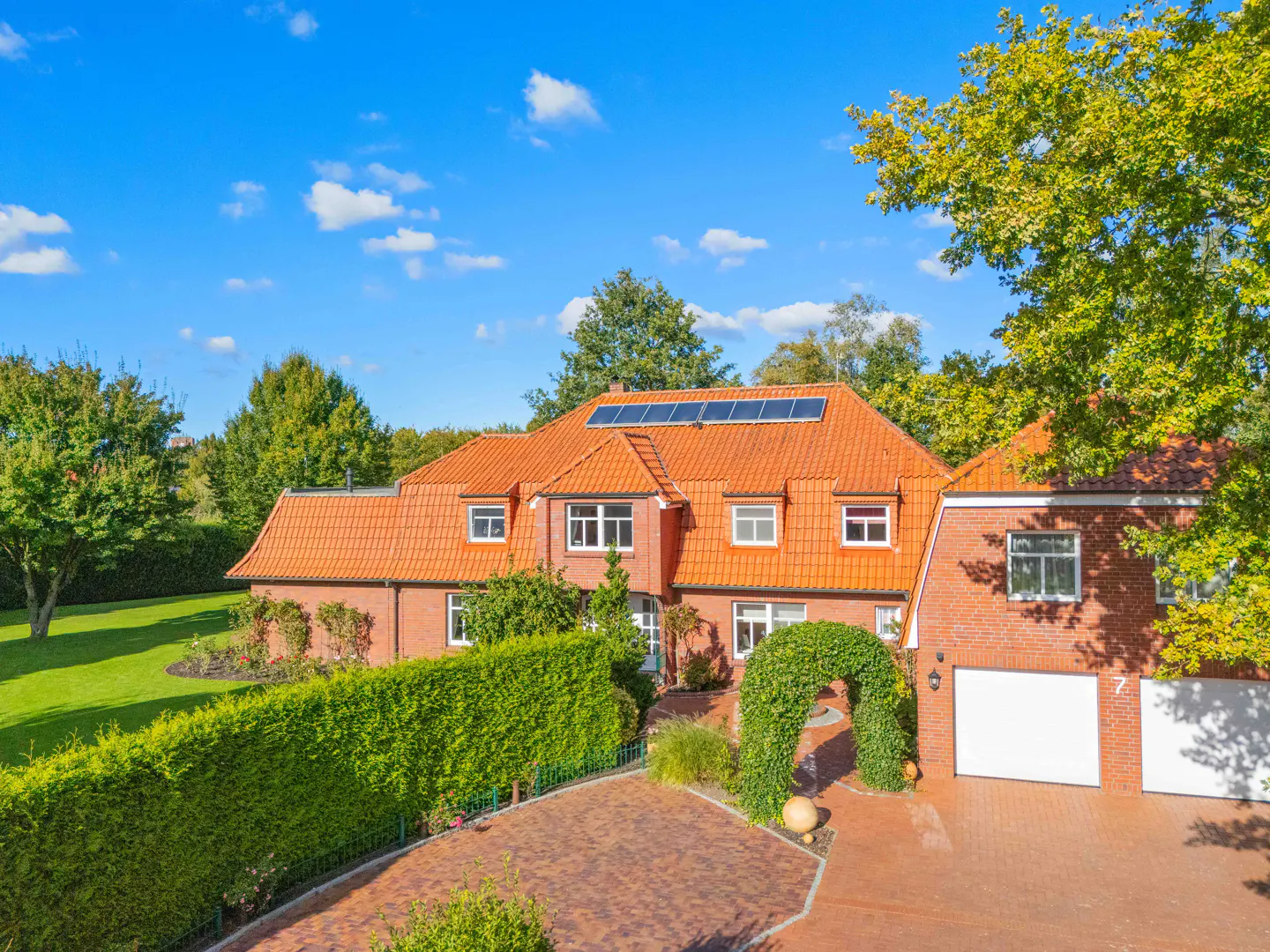 Brick house with orange roof, solar panels, and white garage doors. Green lawn, trees, and blue sky.