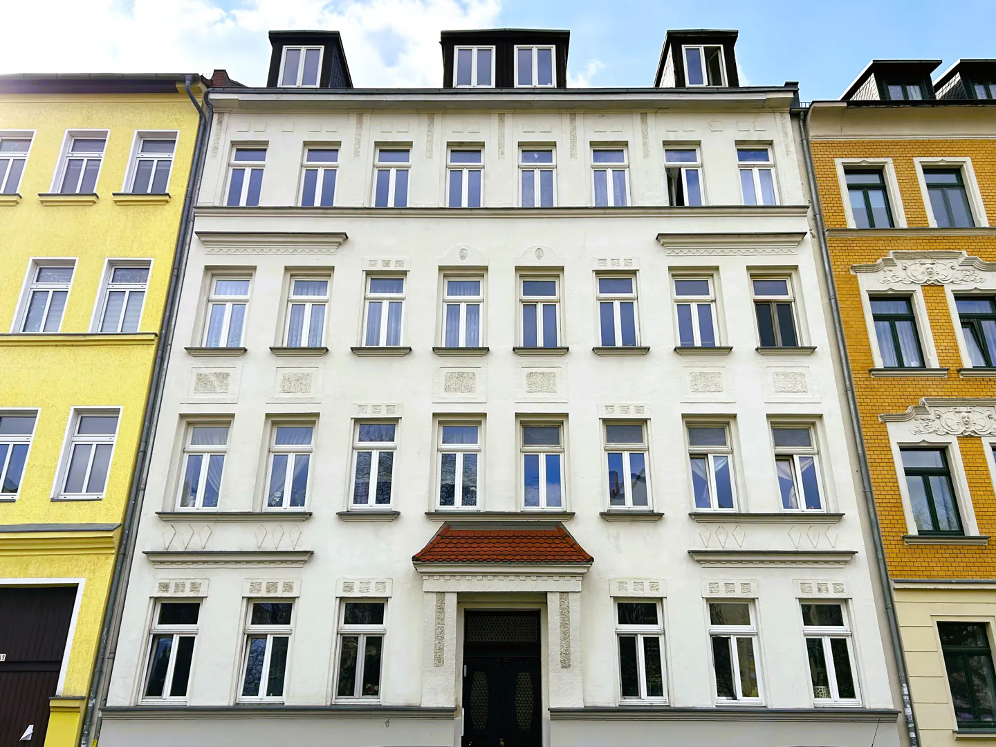 A four-story white building with many windows, flanked by yellow buildings under a blue sky.