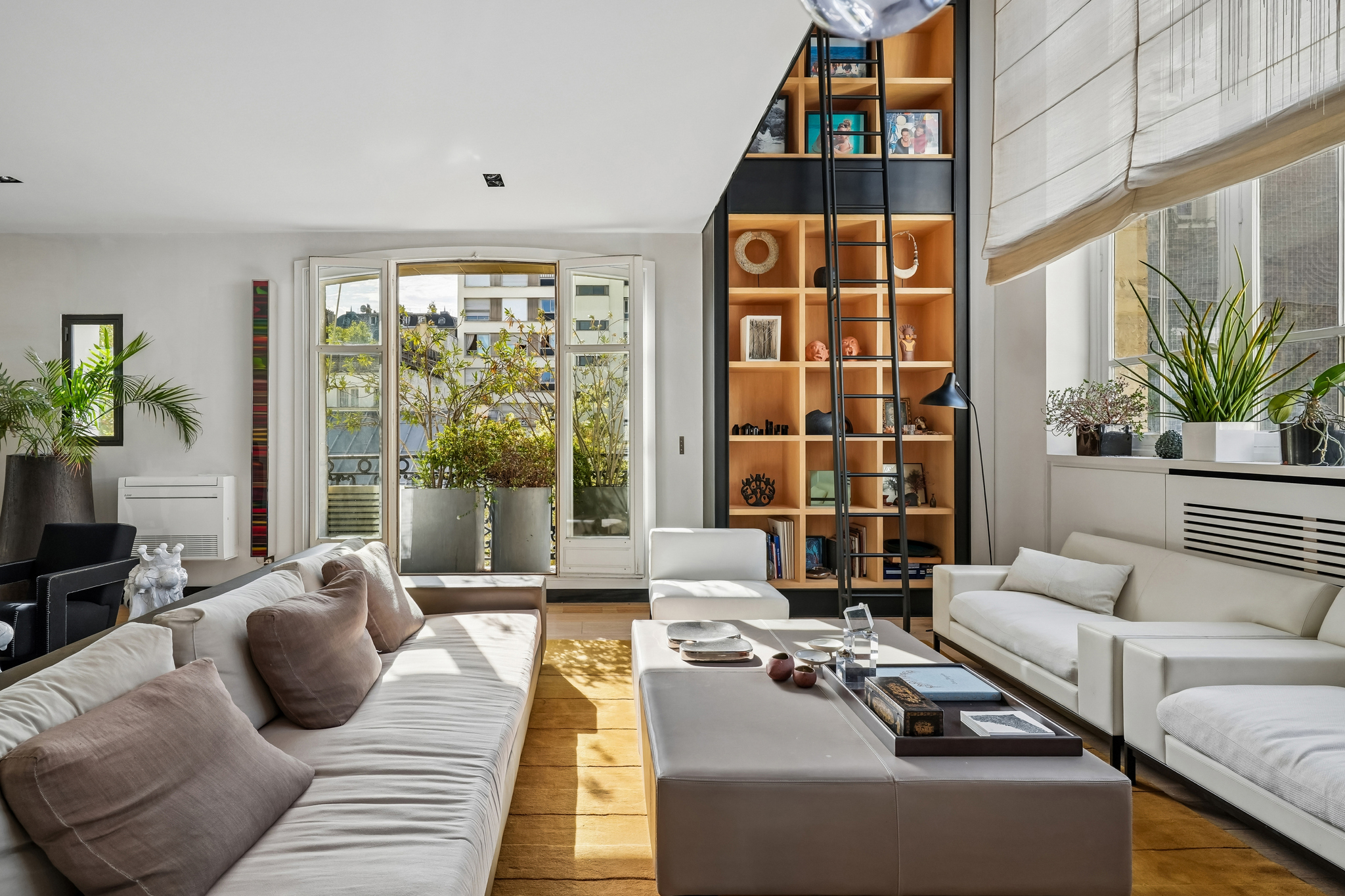 Modern living room with white sofas, a large ottoman, a tall wooden bookshelf with a ladder, and a balcony view. [AI-generated text]