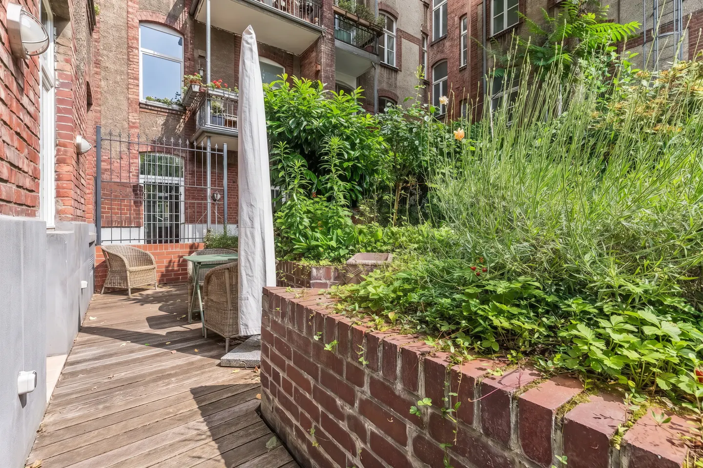 A wooden patio with wicker chairs and a closed umbrella next to a brick planter filled with lush green plants.