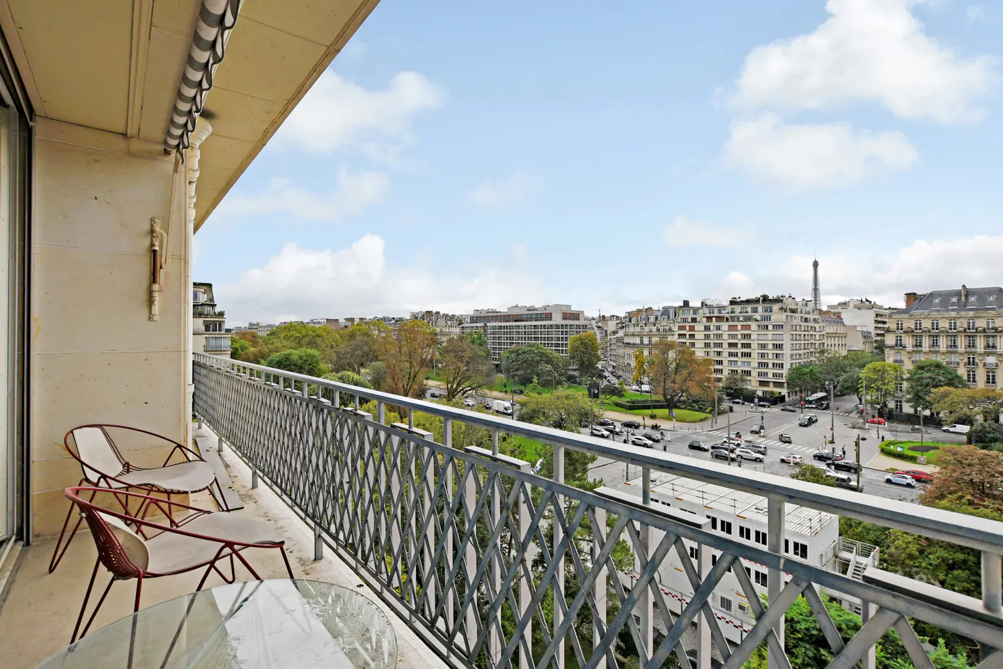 Balcony view of Paris with Eiffel Tower in distance. Two chairs and a table sit on the balcony with a decorative metal railing.