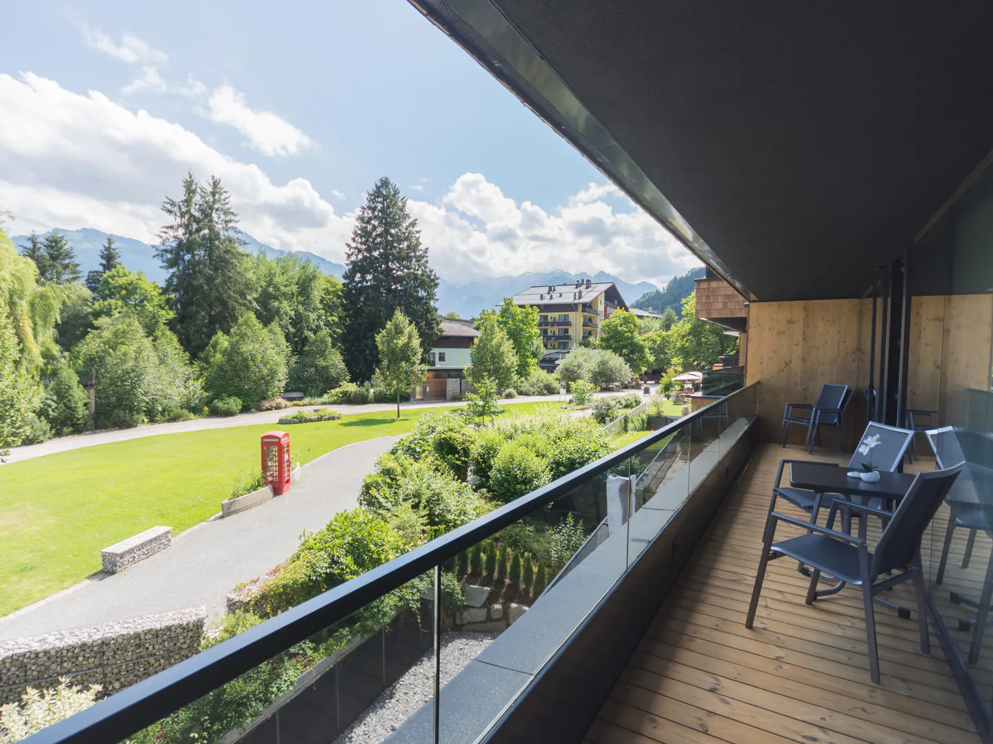 Balcony view of a green lawn with trees, a red phone booth, and mountains in the background. Chairs and a table are on the wooden balcony.