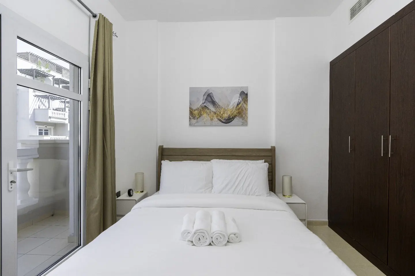 Bright bedroom with white linens, rolled towels, and a wooden headboard. Balcony access with a view, and a dark wood wardrobe.