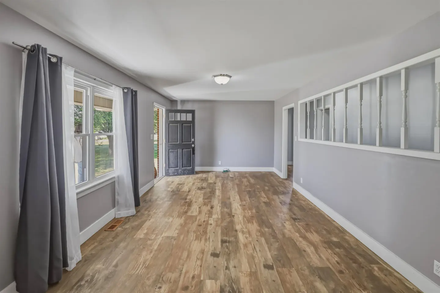A bright, empty living room with gray walls, wood floors, and an open black front door.