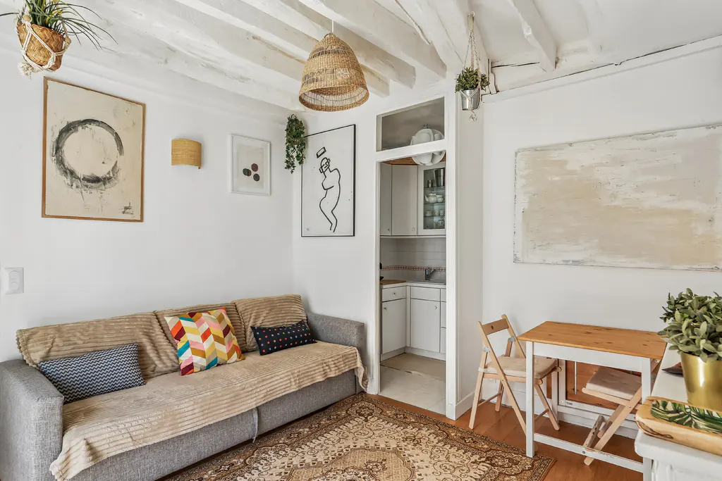 Cozy living room with white walls, exposed beams, and a gray sofa with colorful pillows. A small kitchen is visible through an open doorway.