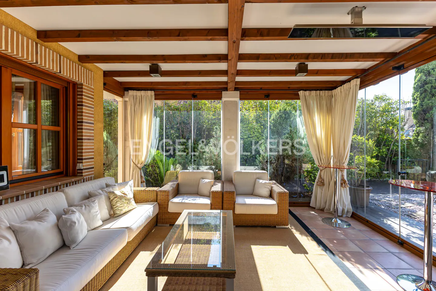 Bright sunroom with wicker furniture, white sofa, glass walls, and wooden beams. Outdoor greenery visible through the glass.
