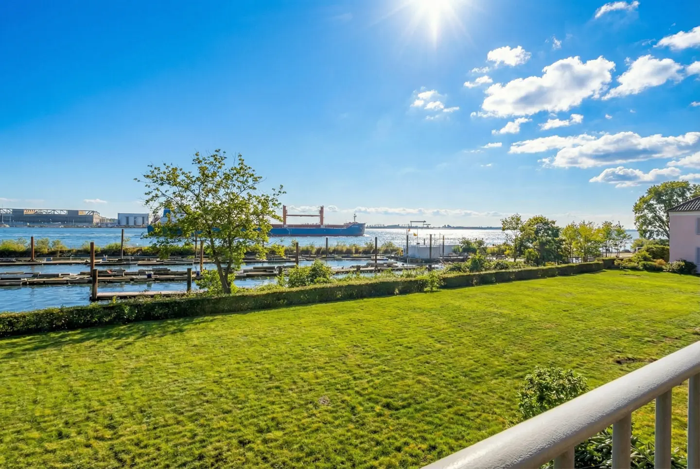 Waterfront view of a green lawn, a tree, and a cargo ship on a sunny day with a blue sky and white clouds.